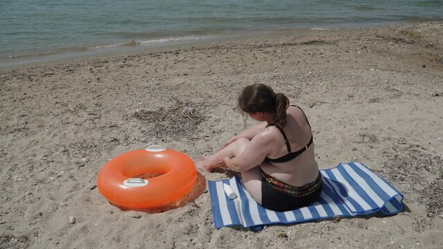 Nordic woman applying sunscreen moisturizer while traveling solo, wide shot. Plus size female person sits on beach blanket on seashore. Ldy takes care of protecting pale sensitive skin.