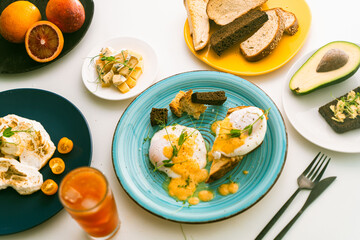 Eggs Benedict with hollandaise sauce and peas on a turquoise plate and avocado with rye toast, cheese, blood oranges and orange juice on a white table.
