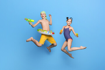 Obraz premium Happy brother and sister in swimwear jumping with water guns on blue studio background