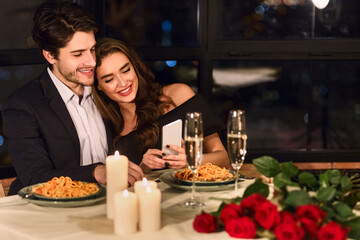 Happy European young couple looking photos on smartphone during romantic dating in restaurant, checking photos