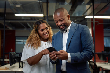 Two happy multiracial business people in businesswear using mobile phone in office