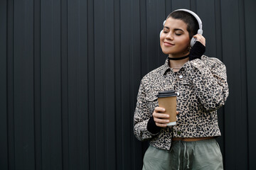 Short-haired woman in chic outfit enjoys coffee while listening to music on headphones near modern...
