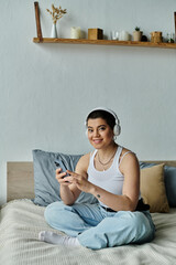 A young woman with short hair sits on a bed, absorbed in her phone screen.