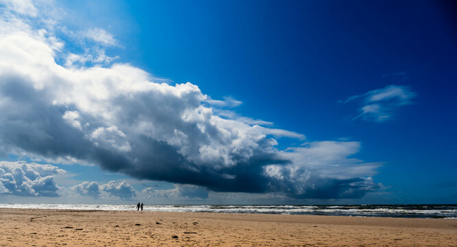 Ein nicht erkennbares Paar geht am hellen Nordseestrand spazieren, im Himmel zeigt sich eine spektakul&auml;r geformte Wolke 