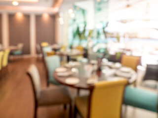 Abstract blur luxury dining room in resort, decoration with wooden furniture, round table and chairs near huge glass window wall with sunlight. Blurred scene of hotel restaurant interior design.