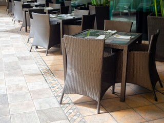 Brown rattan glass dining table with napkins wrapped spoon and fork on the plate mats, empty chair seats, table set preparing for the guests in the resort hotel with nobody, outdoor area.