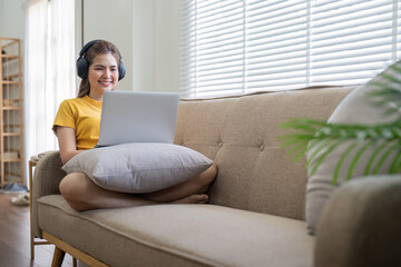 Young Woman Enjoying Music at Home with Headphones While Using Laptop on Sofa in Bright Modern Living Room