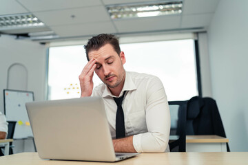 A man is sitting at a desk with a laptop open in front of him. He is wearing a black tie and he is in a state of distress