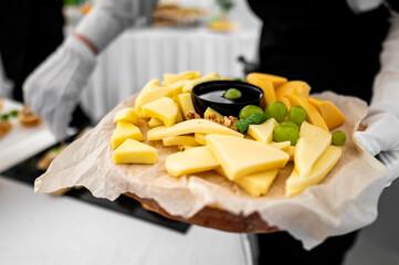 Close-up of a cheese platter with various cheeses, grapes, and nuts on a wooden board. Served by a person in white gloves against a blurred background