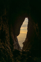 A small opening in a rock wall with the ocean in the background. The sky is a mix of orange and...