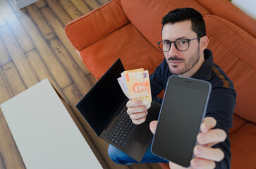 Hombre joven emprendedor generando ventas y dinero online con su móvil y portátil desde casa
