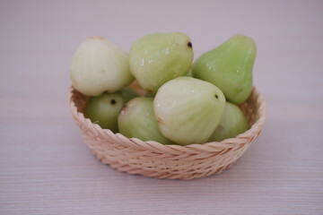 A small wicker basket filled with light green wax apples sits on a pale wooden surface. The fruit has a glossy, slightly bumpy texture and a conical shape.