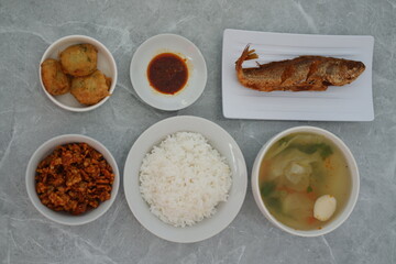 A meal consisting of a plate of fried fish, a bowl of white rice, a bowl of vegetable soup with meatballs, a small plate of red sauce and others, all on a gray marble surface.