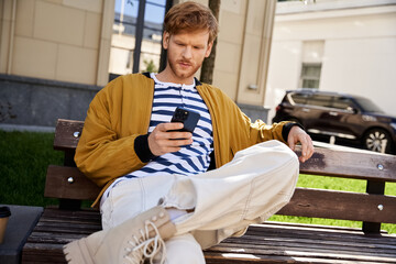 Young man in debonair attire, red hair, sitting on bench, engrossed in cell phone.