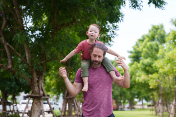 Man with child on shoulders in park, kid extends arms like airplane, both enjoying sunny day outdoors. Relaxation day of Caucasian father and son on the weekend