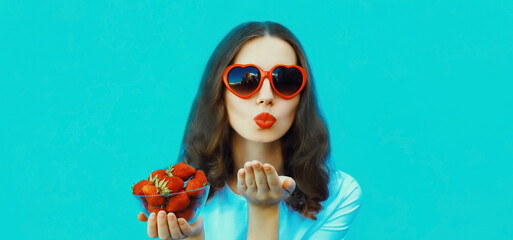 Portrait of beautiful young woman with fresh strawberries blowing a kiss in red heart shaped glasses