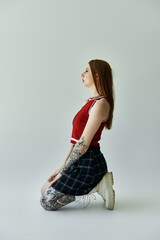 A young woman with tattoos on her arms and legs poses in a plaid skirt and red tank top against a...