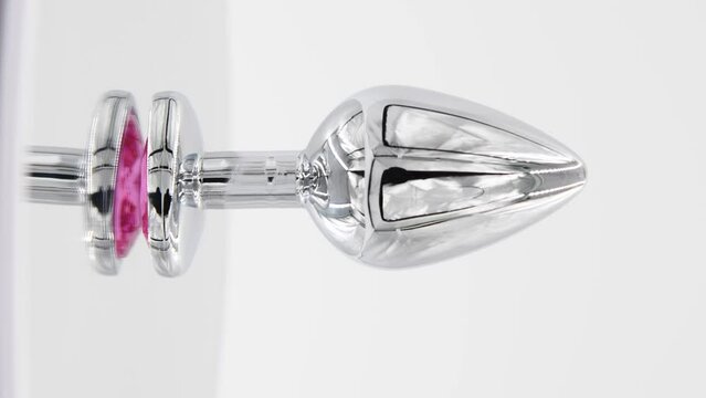 Close-up side view of metallic butt plug with pink heart shaped crystal spinning on mirror surface against white background. Soft focus. Real time vertical video. Adult sex toys theme.