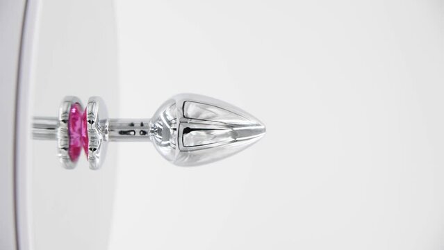 Close-up side view of metallic butt plug with pink heart shaped crystal spinning on mirror surface against white background. Soft focus. Real time vertical video. Adult sex toys theme.