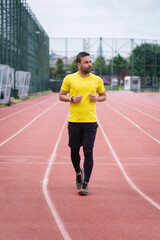 Obraz premium Vibrant young man in a bright t-shirt gracefully runs on an empty track with a red rubber surface and white markings at an outdoor sports ground, healthy lifestyle. 
