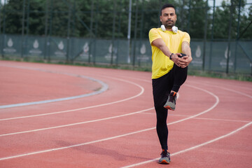 Obraz premium Dynamic runner with wireless headphones limbers up leg muscles on rubber running track at urban park's sports field. 