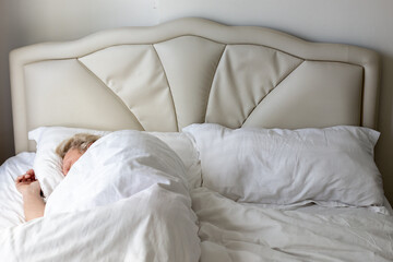blonde mature man sleeps on a bed with a white headboard under a white blanket in the bedroom. Comfortable sleep.