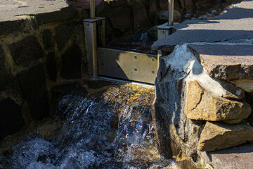 Flowing water in a man-made stone trough. Water flowing over stones. Strong stream of water, rocky bed. A small stainless steel sluice gate, dam.