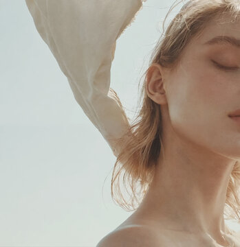 The image is a close-up of a woman's face in profile, with her eyes closed. She has light colored hair and is wearing a cream-colored silk scarf that is blowing in the wind.


