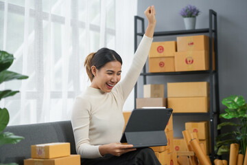 Woman celebrating success while working on a tablet, surrounded by boxes, symbolizing online...