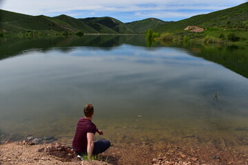 Fototapeta premium Young athletic man in a bordeaux t-shirt crouching at the shore of Montpellier Reservoir and looking at the still water