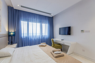 The bedroom suite in the hotel is in light colors with blue curtains on the window.