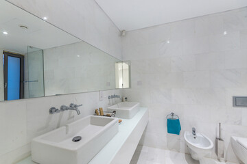The hotel bathroom has white tiles on the walls and floor with a large mirror and two sinks..