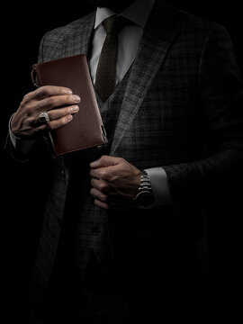 Stylish bearded black gentleman in well-fitting three-piece suit and wristwatch holding sophisticated black leather wallet, embodying understated elegance, standing against dark background.