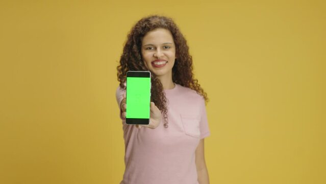 Woman hold smartphone with green screen chroma key show thumb up mock up recommend good application promotional sale offer, point to phone screen. Girl isolated on yellow
