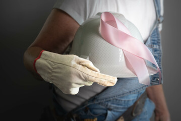 Unrecognizable, female, construction worker wearing blue jean, denim, overalls, holding white hard hat with a pink, breast cancer awareness ribbon
