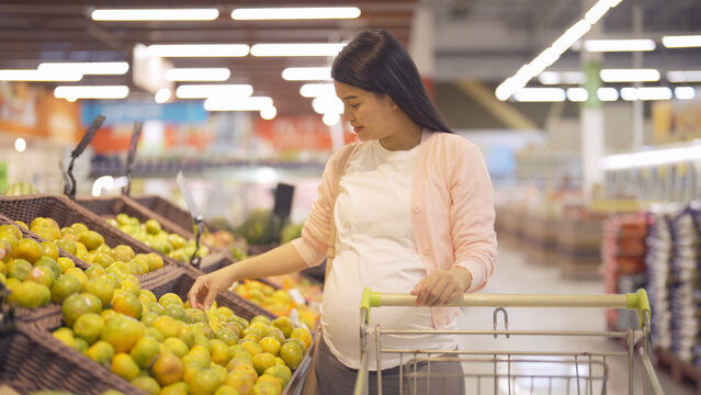 A portrait of happy Asian pregnant woman customer walking along supermarket, buying items on grocery products shelves with basket. Food shopping. Having a baby. Family people lifestyle. Mom.