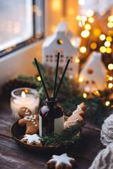 Xmas spirit, simple way to creating holiday atmosphere and festive mood at home on the window sill. Scented burning aromatic candles on wooden tray, liquid reed diffuser with sticks, fir tree branch