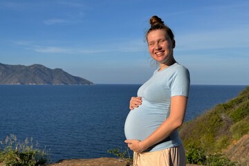 Young pregnant woman stroking her belly outdoors on sea, beach at summer vacation 