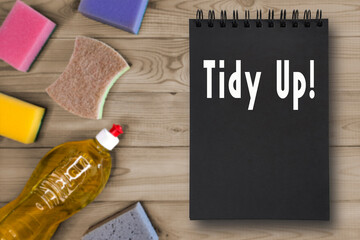 Tidy up lettering. Notebook with black pages lies on wooden table next to washing up liquid in bottle and colorful kitchen sponges.