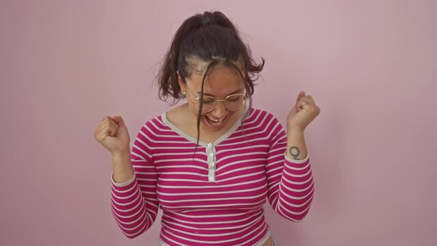 Ecstatic young hispanic woman in stripes t-shirt madly celebrates a triumphant win, standing victoriously with raised arms, eyes closed in joy over pink isolated background.