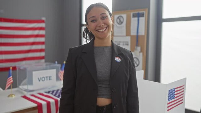 Smiling hispanic woman with 'i voted' sticker pointing at camera in voting room with american flags
