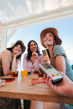 Vertical. Happy young group of only women paying the bill with a contactless credit card in cocktail beach bar. Female smiling holding a creditcard and giving a payment transaction to the cashier