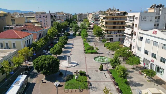 An aerial view of the city center of Kalamata, Greece, showcasing a pedestrian-friendly promenade lined with trees, buildings, and a central fountain.