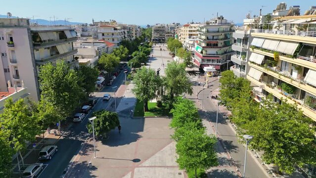 An aerial view of the city center of Kalamata, Greece, showcasing a pedestrian-friendly promenade lined with trees, buildings, and a central fountain.