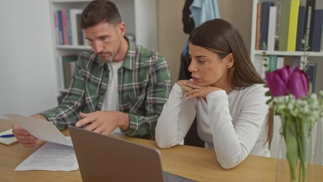 A concerned man and woman review documents and a laptop in a home setting with a bouquet of tulips.