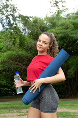 Yogist woman happily smiling holding yoga mat and water bottle. Getting ready for stretching or workout fitness session in a public park