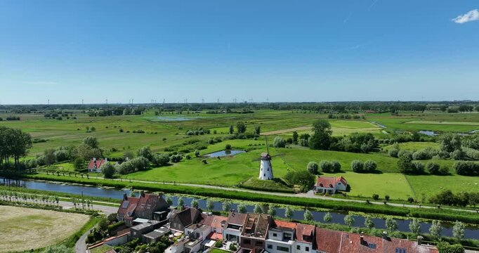Damme, Belgium, June 2022. Drone orbits the Damse Vaart canal with the Schellemolen white windmill, houses, and lush green fields on a sunny day.
