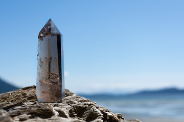 An image of a tall sparkling smoky quartz crystal tower resting on driftwood with the Pacific Ocean in the background. 