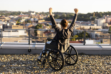 Obraz premium Joyful man in wheelchair raising arms in celebration on a rooftop with cityscape view during sunset.