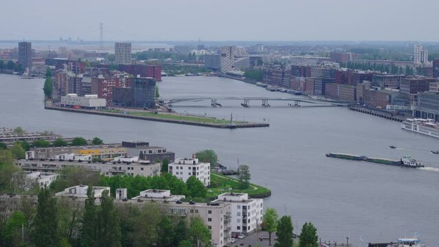 Aerial view of a tanker ship and other boats in Amsterdam's IJ river, framed by a variety of buildings on all sides, on a hazy spring day